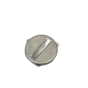 Può includere: Un coperchio circolare per batteria in metallo color argento con una fessura incassata per cacciavite a taglio e un piccolo segno più in rilievo. Il componente presenta tre piccole linguette che sporgono dal bordo esterno del telaio.