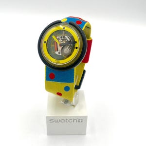 May include: A Swatch watch with a yellow face and a black bezel. The watch band is yellow with blue and red polka dots. The watch is displayed on a white stand with the word "swatch" printed on it.