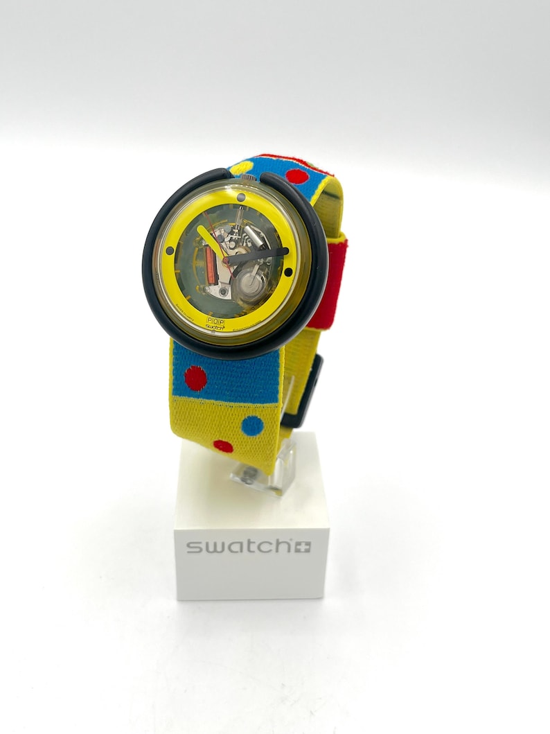 May include: A Swatch watch with a yellow face and a black bezel. The watch band is yellow with blue and red polka dots. The watch is displayed on a white stand with the word "swatch" printed on it.