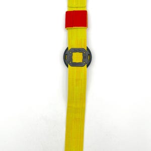 May include: A yellow strap with a black buckle and a red patch. The strap has a black square-shaped component in the middle. The item is designed for securing or adjusting.