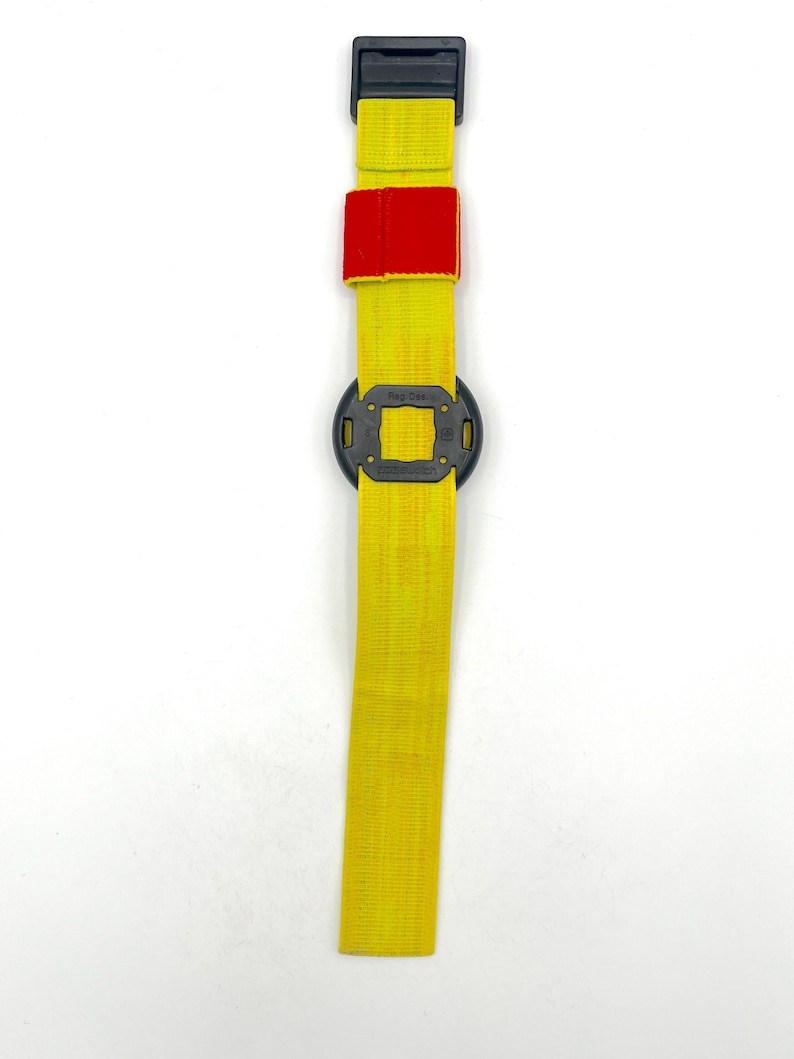 May include: A yellow strap with a black buckle and a red patch. The strap has a black square-shaped component in the middle. The item is designed for securing or adjusting.