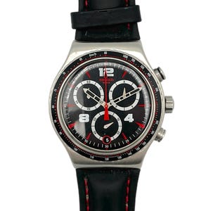 Op de afbeelding: Een zwarte en zilveren Swatch chronograaf horloge met een zwarte leren band en rode stiksels. De wijzerplaat is zwart met witte en rode accenten, waaronder de cijfers 12, 8, 4 en 5. Het horloge heeft drie sub-wijzerplaten.