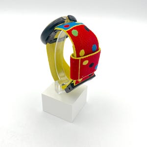 May include: A black smartwatch with a yellow band and a red strap featuring colorful polka dots. The watch is displayed on a white block, showcasing its vibrant design and playful aesthetic. The watch band is approximately 1 inch wide.