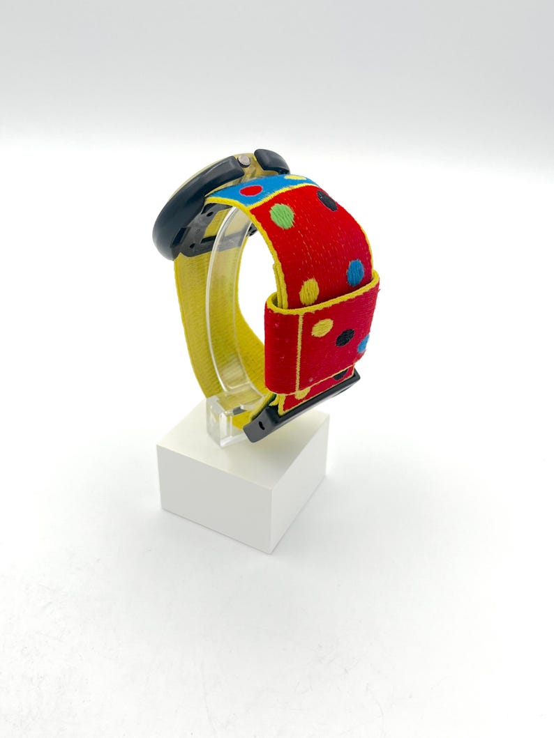 May include: A black smartwatch with a yellow band and a red strap featuring colorful polka dots. The watch is displayed on a white block, showcasing its vibrant design and playful aesthetic. The watch band is approximately 1 inch wide.