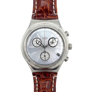 May include: A silver Swatch chronograph watch with a light-colored face and three sub-dials. The watch has a brown leather band with white stitching. The date is displayed at the 3 o'clock position.