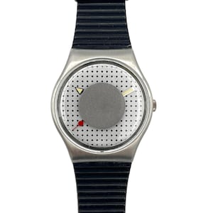 Könnte beinhalten: Eine silberfarbene Armbanduhr mit einem schwarzen, gerippten Armband. Das Zifferblatt hat einen grauen Kreis in der Mitte, ein gepunktetes Muster und einen kleinen roten Punkt. Die Zeiger sind ein gelbes Dreieck und eine dünne weiße Linie.
