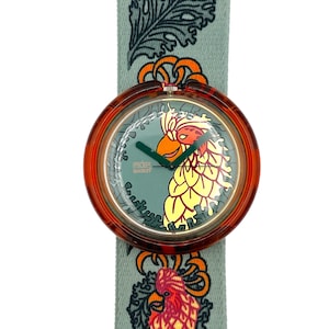 Op de afbeelding: Een ronde horloge met een bruine kast en een groene wijzerplaat met een papegaai-ontwerp. De horlogeband is lichtgroen met een herhalend patroon van veren en papegaaien. De wijzerplaat leest "POP Swatch".