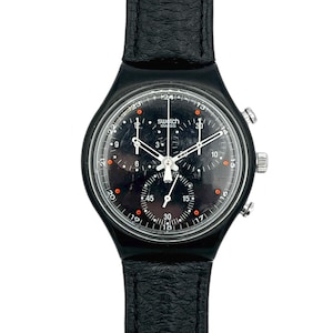 Può includere: Orologio cronografo Swatch nero con cinturino in pelle nera. Il quadrante dell'orologio è nero con dettagli bianchi e arancioni, tra cui la scritta "Swatch" e "Swiss". L'orologio ha tre sotto-quadranti e un datario.