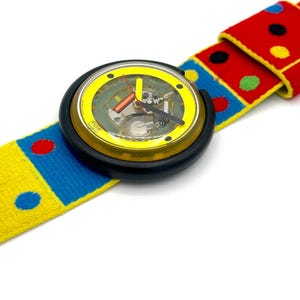 May include: A colorful wristwatch with a yellow face and black casing. The watchband is a vibrant mix of red, blue, and yellow with polka dots. The watch hands are red and black. The watch face has the word "POP" printed on it.