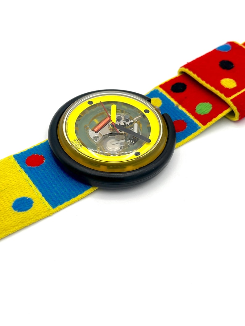 May include: A colorful wristwatch with a yellow face and black casing. The watchband is a vibrant mix of red, blue, and yellow with polka dots. The watch hands are red and black. The watch face has the word "POP" printed on it.
