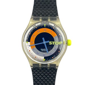 Op de afbeelding: Een Swatch-horloge met een doorzichtige plastic behuizing, een zwarte getextureerde band en een rode en oranje accent. De wijzerplaat is donkerblauw met een witte en oranje ring, en het woord "STOP" is erop gedrukt.