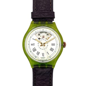 Op de afbeelding: Een Swatch horloge met een transparante groene kast en een witte wijzerplaat met Romeinse cijfers. Het horloge heeft goudkleurige wijzers en een bruine leren band. De wijzerplaat toont de tekst "automatic" en "Swatch AG 1991".
