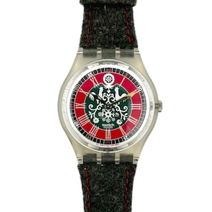May include: A Swatch watch with a clear plastic case and a dark green fabric band with red stitching. The watch face features a red and green design with white accents and the word "Swatch" in white.