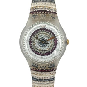 Op de afbeelding: Een Swatch-horloge met een transparante plastic kast en een patroonband. De wijzerplaat heeft een cirkelvormig ontwerp met witte, paarse en bruine elementen. De band heeft een bijpassend patroon. Het horloge heeft goudkleurige accenten.