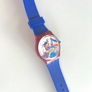 Peut inclure: Une montre-bracelet bleue et rouge avec un cadran graphique repr&eacute;sentant un homme et un drapeau. La montre a un bracelet en silicone bleu. Le cadran de la montre est rouge et le bracelet est bleu.