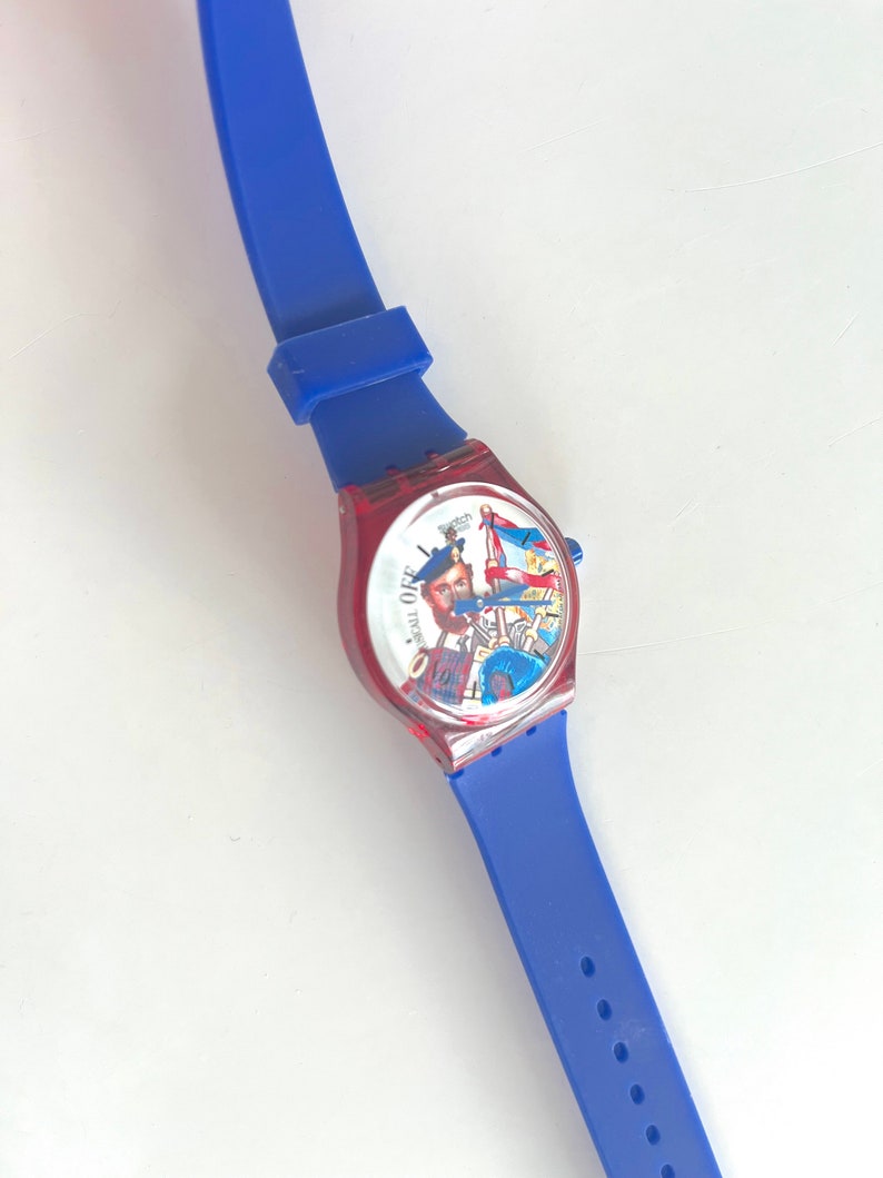 Peut inclure: Une montre-bracelet bleue et rouge avec un cadran graphique repr&eacute;sentant un homme et un drapeau. La montre a un bracelet en silicone bleu. Le cadran de la montre est rouge et le bracelet est bleu.