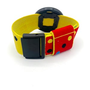 May include: A colorful watch with a yellow strap and a black watch face. The strap features a red section with polka dots and a black buckle. The watch is designed with a playful and vibrant aesthetic.