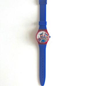 Peut inclure: Une montre Swatch bleue avec un bracelet assorti. Le cadran de la montre est rouge avec une illustration color&eacute;e d'un homme en kilt jouant de la cornemuse. Le bracelet mesure environ 23 cm de long.