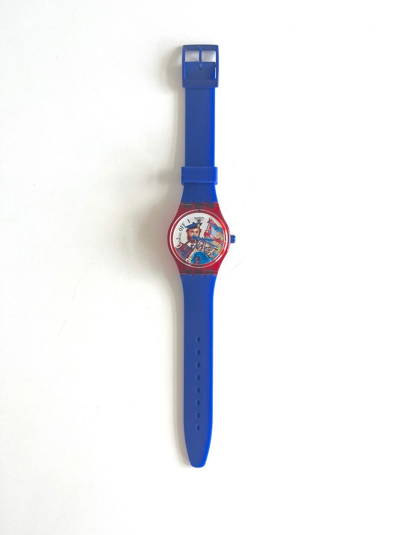 Peut inclure: Une montre Swatch bleue avec un bracelet assorti. Le cadran de la montre est rouge avec une illustration color&eacute;e d'un homme en kilt jouant de la cornemuse. Le bracelet mesure environ 23 cm de long.