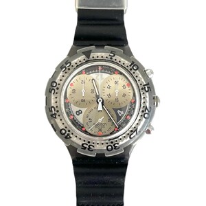 May include: A silver-toned Swatch watch with a black rubber strap. The watch face has a light beige background with black and red accents, and three sub-dials. The watch features the text "Swatch Swiss 30".