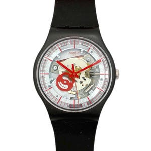 May include: A black Swatch watch with a transparent face revealing the internal mechanisms. The watch features red hands and accents, a white dial with red markings, and a red strap. The watch is a stylish accessory.