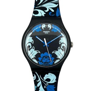 Pode incluir: Um relógio de pulso preto com um padrão floral em tons de azul claro e escuro na bracelete e no mostrador. O mostrador apresenta o texto swatch swiss em branco, com ponteiros azuis e brancos que indicam as horas sobre um fundo preto.