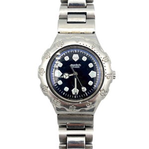 Op de afbeelding: Een zilverkleurig Swatch-horloge met een donkerblauwe wijzerplaat en een bijpassende metalen band. De wijzerplaat heeft witte geometrische uurmarkeringen en het Swatch-logo. De lunette heeft verhoogde markeringen. Het horloge wordt tegen een witte achtergrond getoond.