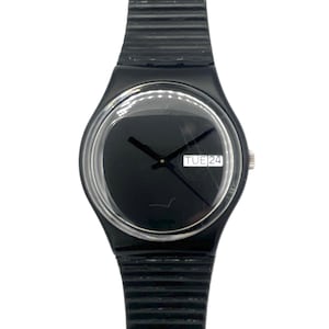 Op de afbeelding: Zwarte polshorloge met een ronde wijzerplaat en een zwarte band. De wijzerplaat toont 'TUE 24'. Het horloge heeft een transparante afdekking en een zilveren kroon. De band is geribbeld.
