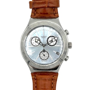 Op de afbeelding: Een zilverkleurig Swatch Swiss chronograaf horloge met een lichtblauwe wijzerplaat en een bruine leren band. Het horloge heeft drie sub-wijzerplaten, een datumvenster en een getextureerde wijzerplaat. De band is een rijke bruine kleur.