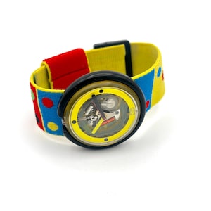May include: A colorful wristwatch with a yellow face and a black bezel. The watchband is a mix of red, yellow, and blue with polka dots. The watch face has visible internal mechanisms. The watch is a fun, retro accessory.