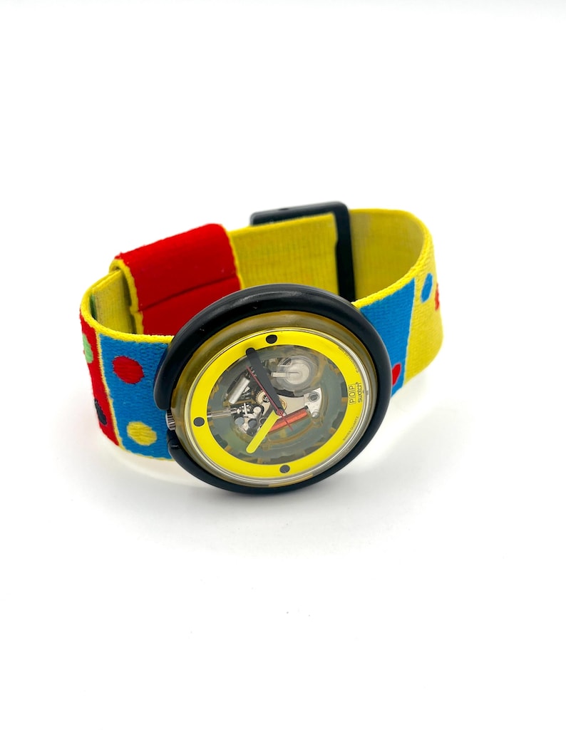 May include: A colorful wristwatch with a yellow face and a black bezel. The watchband is a mix of red, yellow, and blue with polka dots. The watch face has visible internal mechanisms. The watch is a fun, retro accessory.