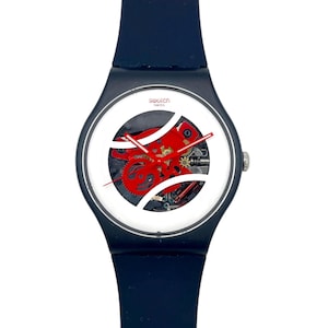 Peut inclure: Une montre Swatch avec un bracelet bleu marine et un cadran blanc. Le cadran a un centre transparent révélant les engrenages internes rouges. Les aiguilles de la montre sont rouges, et la montre est marquée "swatch swiss".
