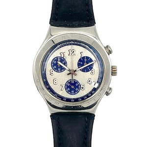 May include: A silver-toned wristwatch with a white face and blue sub-dials. The watch features a black leather band and a silver bezel. The watch face displays the numbers 1 through 12, with a date window at the 4 o'clock position.