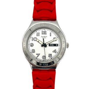 May include: A Swatch Swiss watch featuring a white dial, silver bezel, and a vibrant red silicone strap. The watch face shows black numerals, hands, and the day. A stylish and sporty timepiece with a bold colour scheme.