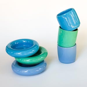 Peut inclure: Un ensemble de vaisselle en céramique composé de trois bols empilés en forme d'anneau et de trois tasses cylindriques empilées. Les objets présentent une finition brillante dans des tons bleu vif, vert éclatant et bleu pervenche.