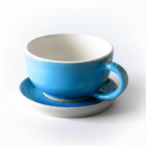 May include: A bright blue ceramic cup with a smooth, glossy finish and a white interior, resting on a matching blue saucer. The cup features a curved handle on the right side, and the entire set is presented against a plain white background.