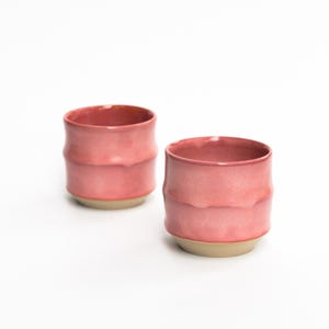 Puede incluir: Dos tazas de cerámica con un esmalte rosa brillante y liso en la parte superior y una base beige mate sin esmaltar. Cada taza presenta una sutil textura horizontal alrededor del centro, creando un perfil suave y ondulado.