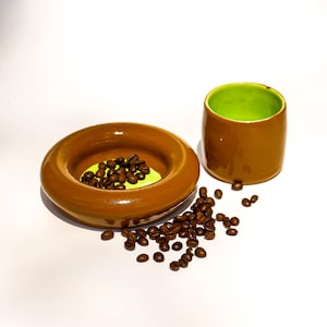 May include: A ceramic cup and a matching circular dish with a glossy brown exterior and a vibrant lime green interior. The dish contains a small pile of roasted coffee beans, with additional coffee beans scattered around the base of the items.