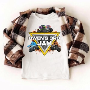 May include: White t-shirt featuring a colorful graphic of monster trucks and the text "OWEN'S 3RD JAM" inside a yellow triangle. A brown and white plaid jacket is draped over the shirt.