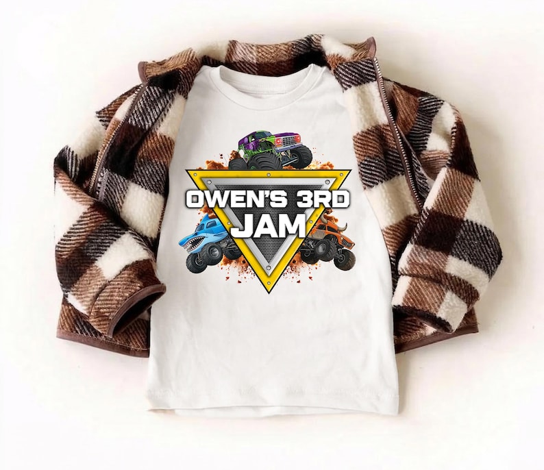 May include: White t-shirt featuring a colorful graphic of monster trucks and the text "OWEN'S 3RD JAM" inside a yellow triangle. A brown and white plaid jacket is draped over the shirt.