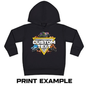Puede incluir: Sudadera con capucha negra con un dise&ntilde;o de cami&oacute;n monstruo colorido. El dise&ntilde;o incluye un tri&aacute;ngulo amarillo con las palabras "CUSTOM TEXT" y tres camiones monstruo. El texto "PRINT EXAMPLE" est&aacute; impreso en la parte inferior.