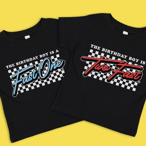 Op de afbeelding: Twee zwarte T-shirts met een geruit racevlagontwerp. Het eerste shirt zegt "The birthday boy is a Fast One" in blauwe tekst. Het tweede shirt zegt "The birthday boy is Two Fast" in rode tekst.