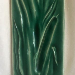 May include: A green ceramic tile with a raised design of a single stalk of grass.