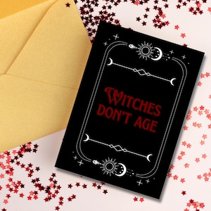 Könnte beinhalten: Schwarze Karte mit rotem Text "Witches Don't Age", umgeben von weißen Himmelsmotiven. Daneben ein goldener Umschlag und rote Sternenkonfetti. Mystisches Thema.