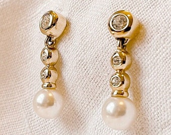 May include: A pair of gold-toned drop earrings. Each earring features a small, round, clear gemstone at the top, followed by three smaller clear gemstones, and a dangling white pearl at the bottom. The earrings are displayed on a white fabric.