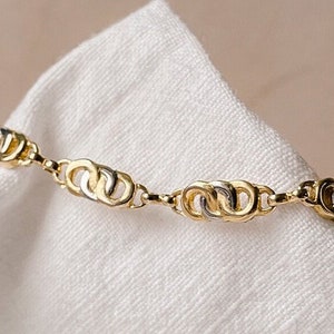 Puede incluir: Un brazalete de oro con una cadena hecha de anillos dobles entrelazados. El brazalete está sobre un fondo de tela blanca.