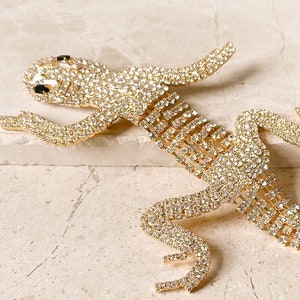 Peut inclure: Une broche en forme de lézard dorée recouverte de strass transparents. Le lézard est en position rampante, la queue enroulée.