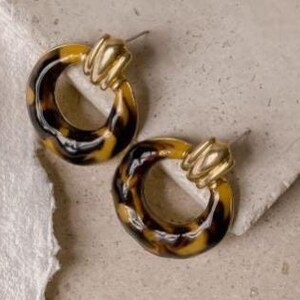 May include: A pair of gold-toned hoop earrings with a tortoiseshell pattern. The earrings have a circular design with a textured gold accent at the top. The earrings are set against a neutral background.