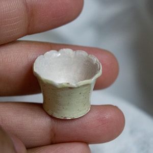 May include: A miniature ceramic bowl with a scalloped rim, featuring a pale yellow exterior speckled with small reddish-brown dots and a smooth, off-white interior. The small vessel has a flared shape and a glossy finish on its surface.