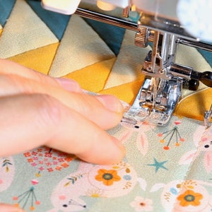 May include: A close-up of a sewing machine needle stitching two pieces of fabric together. The top fabric is a yellow and blue geometric pattern, and the bottom fabric is a green fabric with a white bunny rabbit pattern. The sewing machine foot is a walking foot.